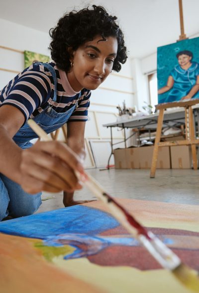 Studio, art and a woman painting on floor in design school with creativity and paint brush. Artist student from India, happy painter and a girl working on creative school project canvas in classroom